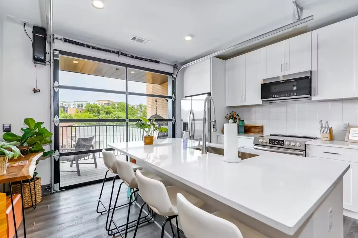 A spacious kitchen interior featuring a large kitchen island, an open garage door leading to outdoor seating, and plenty of room for cooking, entertaining, and casual dining. The scene is bright and cheerful, with clear sightlines from kitchen to outdoors.