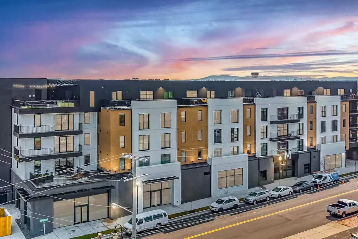 A striking exterior shot of a modern building set against a colorful sunset sky. The architecture features clean lines and large balconies, evoking a vibrant, contemporary feel. Hints of community life are visible, with spaces designed for evening gatherings and relaxing while taking in the neighborhood atmosphere.