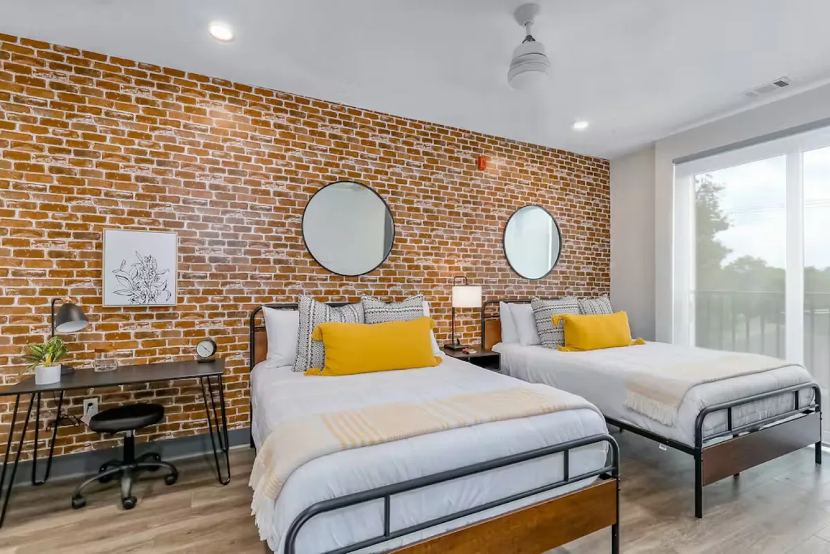 A bright, modern living space featuring a brick accent wall, round mirrors, a yellow accent pillow, and a compact desk with a chair set near the window. The scene suggests a multi-functional area suitable for both work and relaxation, with natural light streaming in and a cozy seating arrangement.