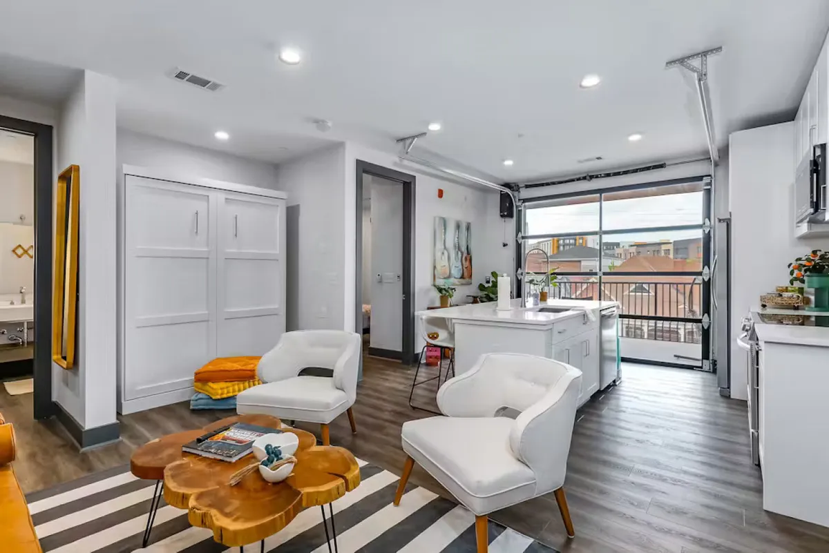 This image showcases a contemporary kitchen and living area with white armchairs, a wooden coffee table, a prominent kitchen island, and large windows letting in abundant natural light. The kitchen island is a central feature, inviting social interaction and meal preparation, while the open layout suggests a seamless flow between cooking and relaxing.