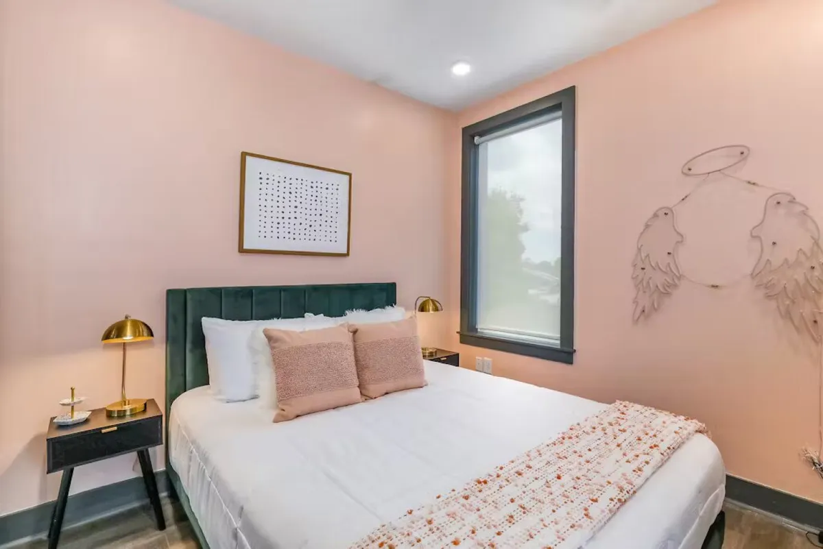 This image showcases a bedroom with a green velvet headboard, decorative wall art, soft pink walls, and gold table lamps. The scene feels serene and cozy, with natural light enhancing the tranquil atmosphere, and plush bedding promoting restful sleep.