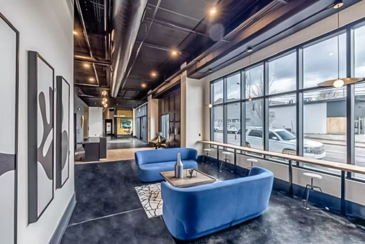 A stylish lounge featuring a blue sofa, large windows providing ample natural light, contemporary wall art, and a bar table. The space is set up for casual meetings, social gatherings, or relaxation, with views that suggest a connection to the surrounding urban environment.