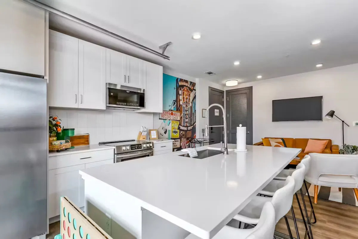 The scene captures a kitchen island with comfortable seating, a colorful wall mural, and a seamless connection to the adjacent living area. The space radiates a casual, welcoming vibe, ideal for cooking, entertaining, and keeping the group together.