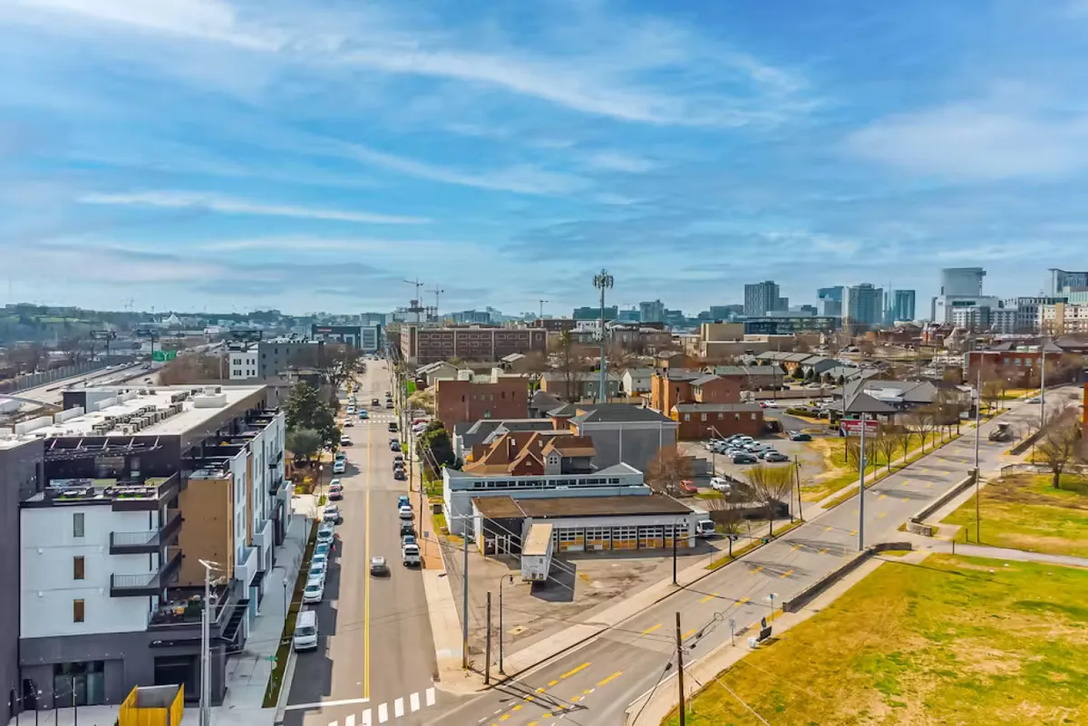 A lively street scene with modern apartment buildings, a tree-lined street, the city skyline in the background, and an open green space. The area is active, suggesting opportunities for walking, biking, and exploring the neighborhood’s parks and attractions.