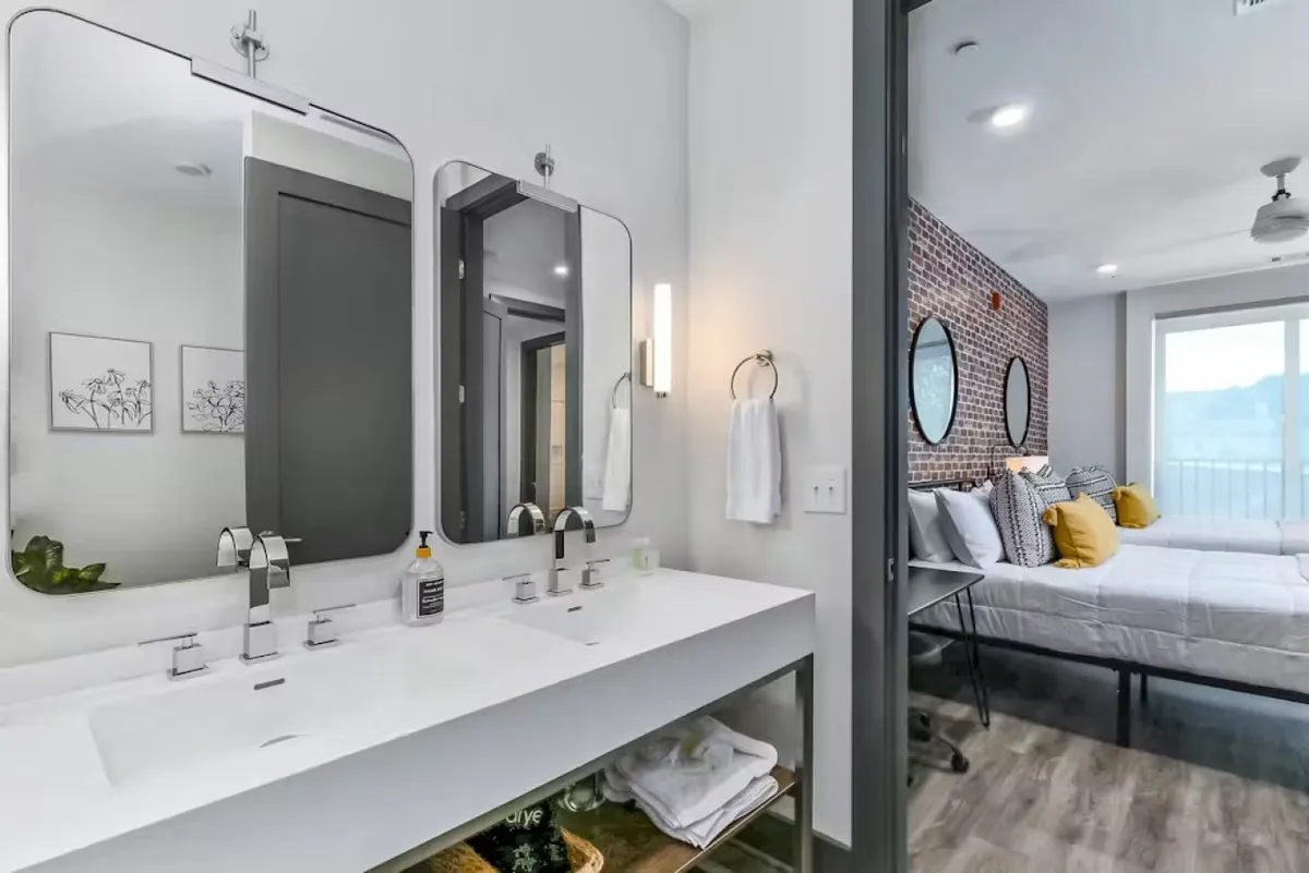 This image features dual sinks with modern faucets, large mirrors, and an exposed brick wall. In the background, comfortable bedding with decorative pillows is visible, tying the bathroom and bedroom together. The mood is fresh and contemporary, with a focus on the spa-inspired, rejuvenating elements.