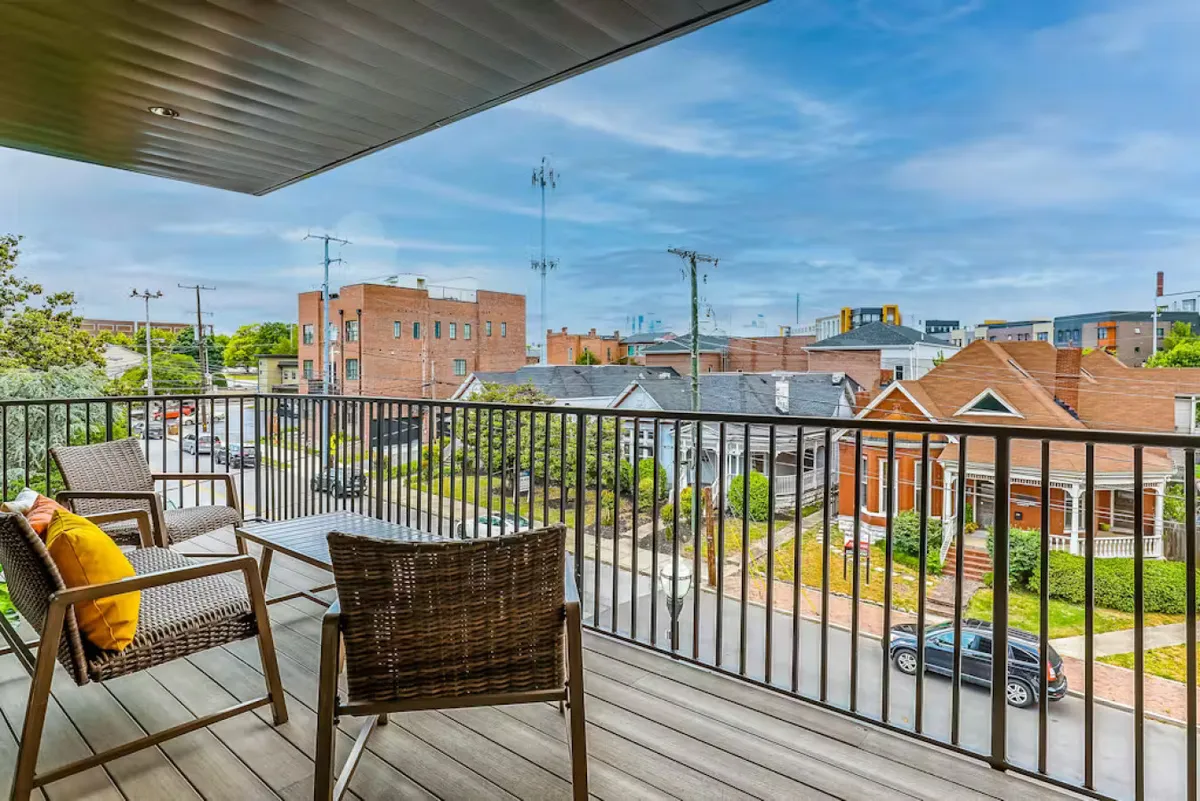 A cozy outdoor balcony with comfortable seating, decorative cushions, and a view over the lively neighborhood. The scene is casual and inviting, perfect for relaxing, sharing stories, or people-watching after a big night out.
