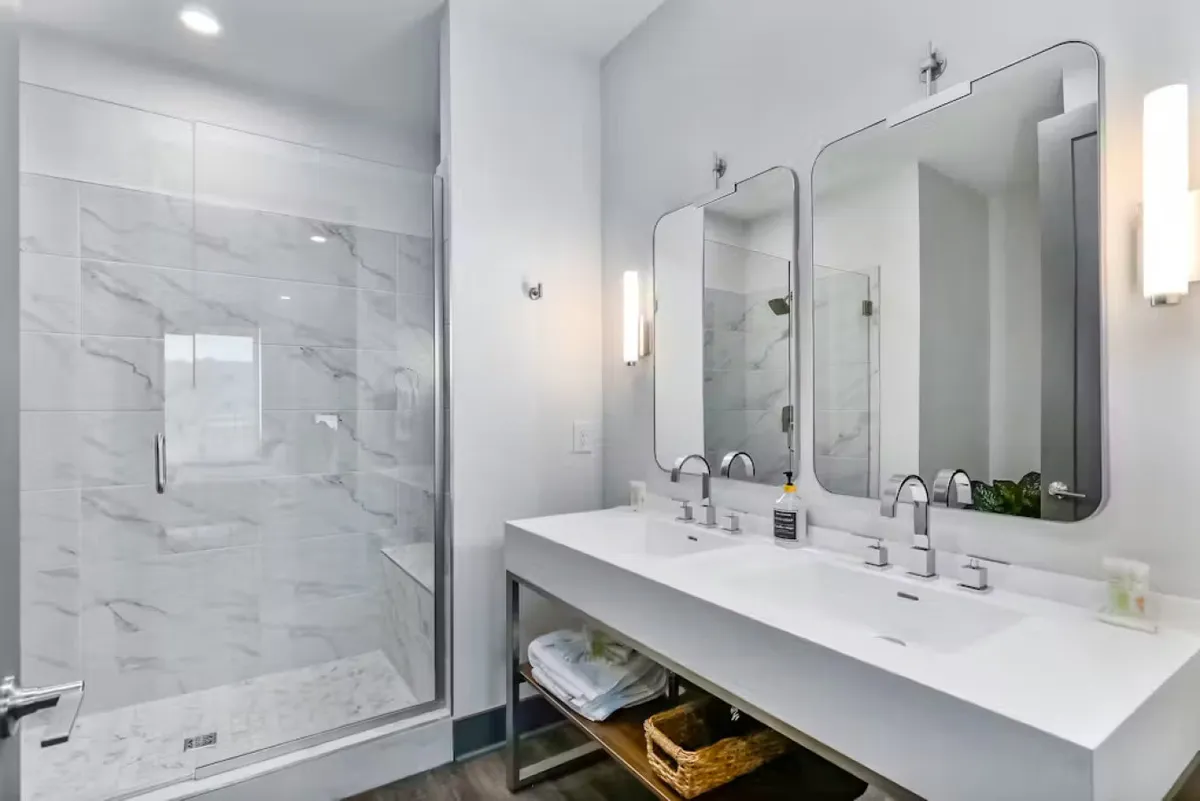 The image shows a double sink vanity with large mirrors and a glass shower enclosure. The space is bright, airy, and exudes a refreshing, serene vibe suitable for self-care rituals and relaxing routines.